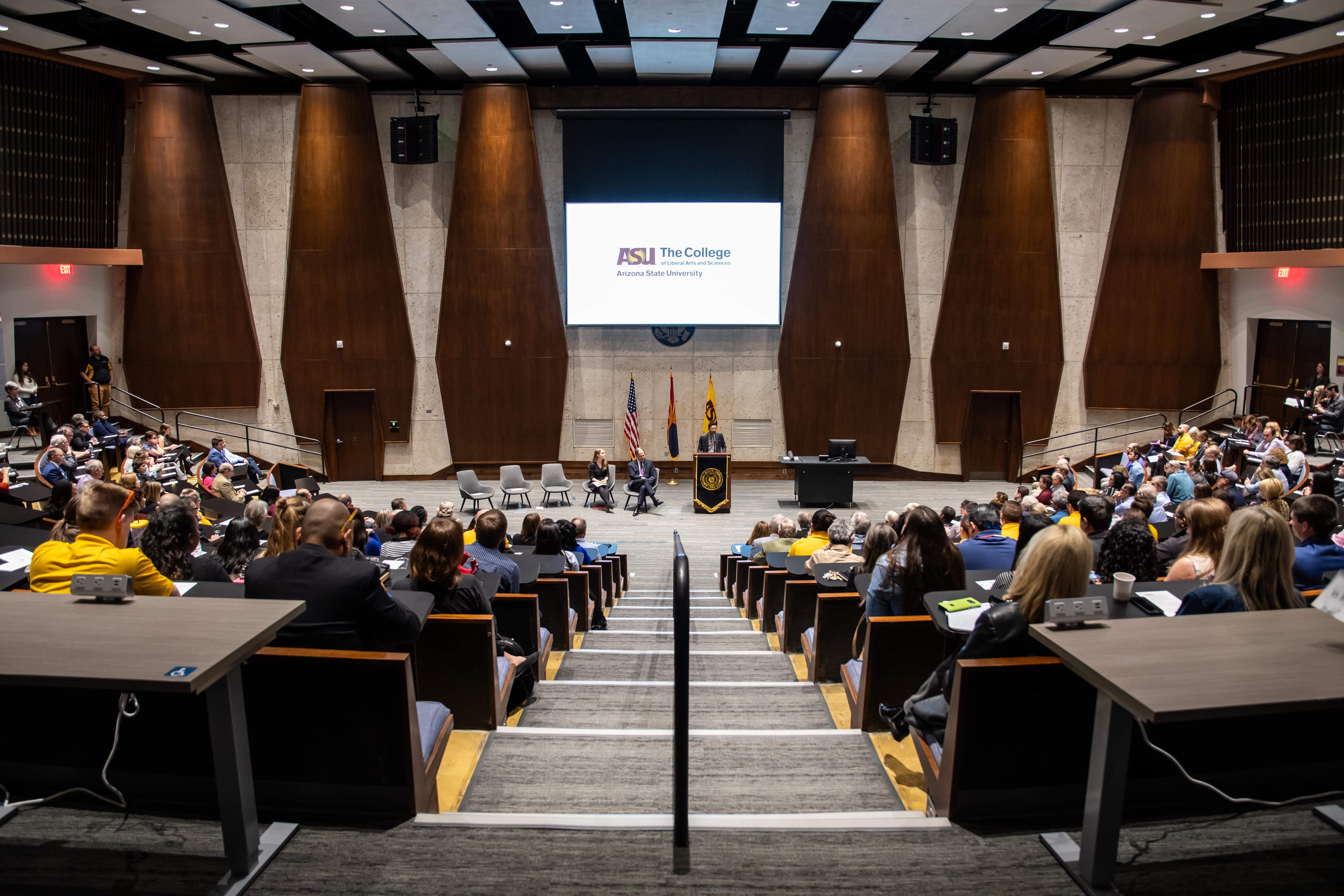 Students sitting in a lecture room with man speaking at podium in front of projected screen